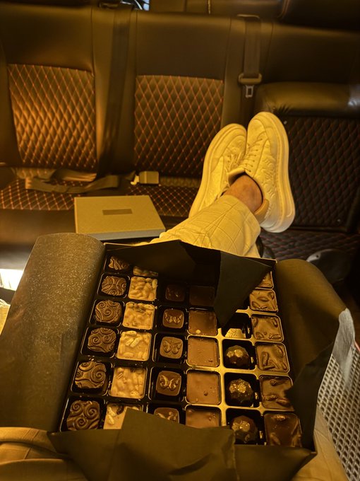 A person's feet rest in white sneakers on a dark leather airplane or vehicle seat, with an open box of assorted chocolate truffles in gold wrappers on their lap.