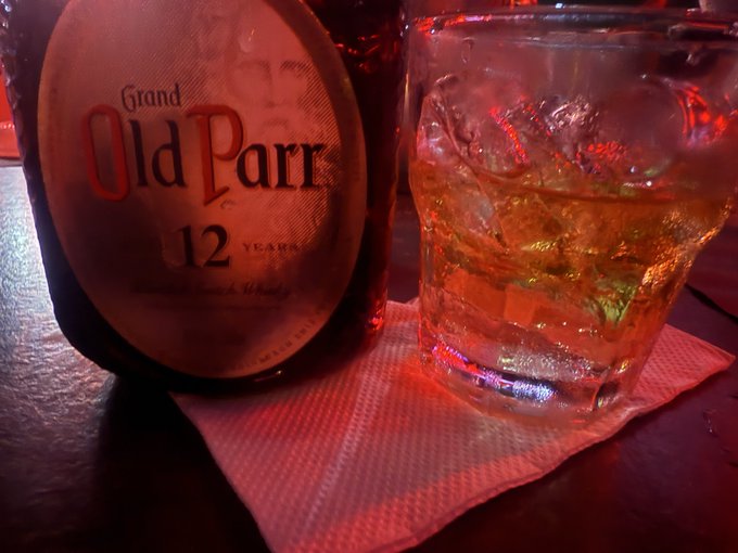 A bottle of "Grand Old Parr 12 Years" whisky sits beside a glass filled with the amber drink over ice, resting on a red cocktail napkin in dim, warm lighting—suggesting an intimate toast to celebrate the couple's 14th anniversary.