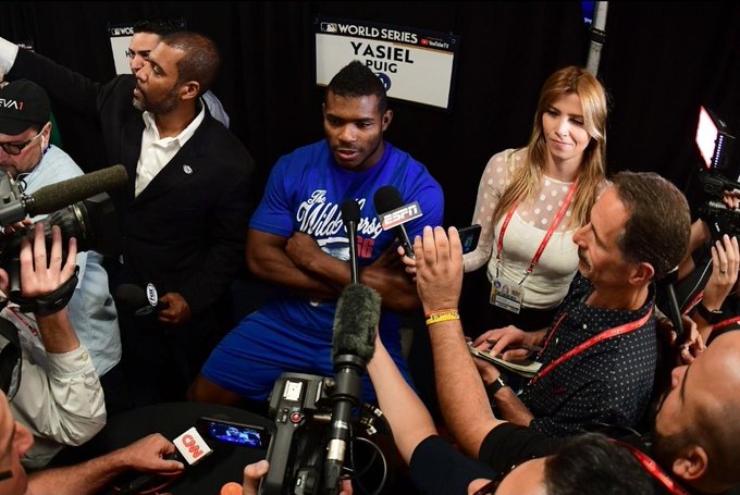 Baseball player Yasiel Puig, wearing a blue 'The White Sox' shirt, is surrounded by reporters and cameras during a World Series media session, with a 'YASIEL PUIG' name placard visible behind him.