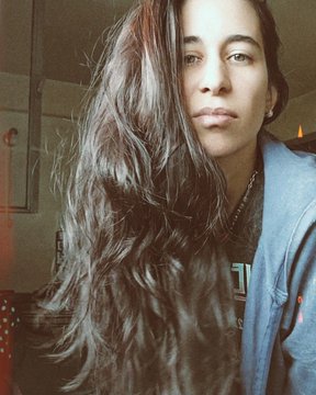 A selfie of a young woman with long wavy brown hair wearing a blue shirt, photographed indoors.