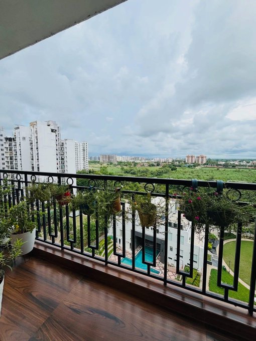 A view from a high-rise balcony decorated with hanging potted plants, overlooking a residential area with white apartment buildings, green fields, and a swimming pool below.