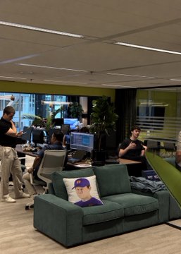 An office workspace featuring a green couch with a baseball-player pillow, with employees working at computers in the background.