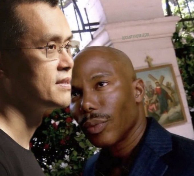 A close-up photo of two men posing closely together, one East Asian and one African American, with a watermark '@CANTPROVEITSOL' visible and a religious painting and floral arrangements in the background.