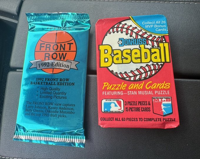 Two unopened vintage trading card packs sit on a surface: a blue '1991 Front Row Basketball Edition' pack and a red 'Donruss Baseball' pack advertising 'Puzzle and Cards' featuring a Stan Musial puzzle.