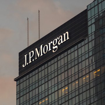The image shows a tall glass office building with "JPMorgan" prominently displayed on its facade against a sunset sky, accompanied by two antennas on top. The scene illustrates the headline about JPMorgan, America's largest bank with $5 trillion in assets, enabling clients to send USD deposits on the Ethereum blockchain using JPM Coin.