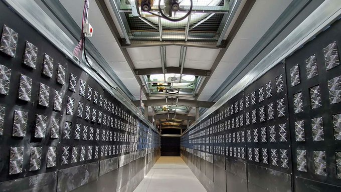 A long industrial corridor lined with rows of silver ventilation or ducting units beneath an exposed ceiling with machinery.