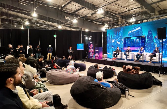 A panel discussion takes place on a stage at 'FUTURLANA' with five speakers seated in front of an audience relaxing on bean bags in a large warehouse-like venue.