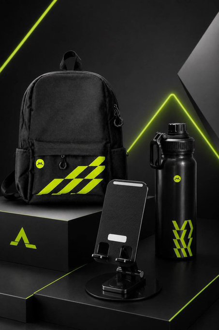 A promotional display showcasing MGBX giveaway prizes: a black backpack with green accents, a black insulated tumbler, and a phone stand, all featuring the MGBX logo. The items are arranged on dark platforms with neon green triangular lighting, matching the branded merchandise described in the giveaway announcement.