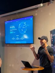 A man in a dark t-shirt and cap gestures while presenting next to a wall-mounted screen showing a blue data dashboard.