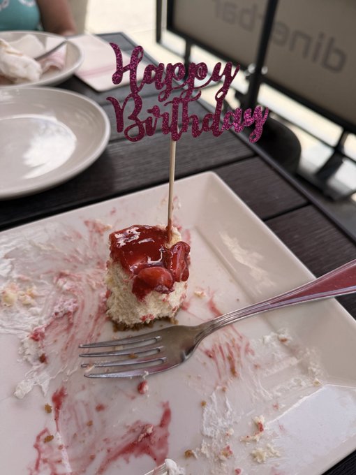 A small square piece of strawberry-topped cake sits on a square white plate, adorned with a sparkly pink 'Happy Birthday' cake topper, with a fork resting on the messy plate.