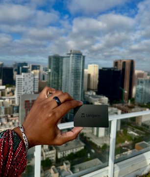 A hand holds a black Tango card against a backdrop of a city skyline on a rooftop, with a red beaded bracelet visible on the wrist.