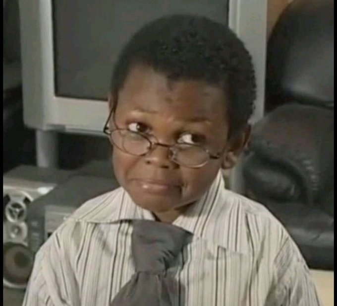 A young boy wearing glasses, a striped dress shirt, and a dark tie gives a skeptical sideways glance while sitting in front of a computer monitor.