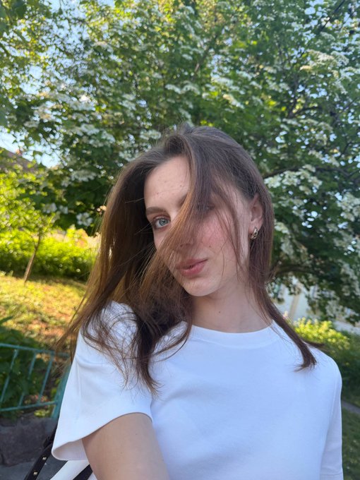 A young woman with long brown hair wearing a white t-shirt poses for a selfie outdoors, with green trees visible in the background.