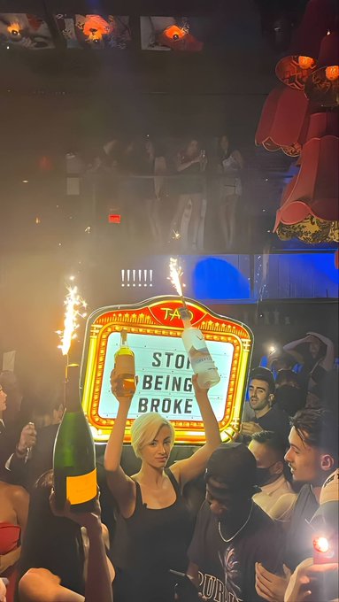 The caption should be:

A lively nightclub scene captures a triumphant woman holding up a bottle and glass in celebration, with a vintage "TAXI" sign displaying "STOP BEING BROKE" illuminated by sparklers in the background, perfectly matching the post's motivational message about ending financial struggles. The festive atmosphere, with crowds cheering and lights flashing, embodies the excitement of a major financial breakthrough ahead.