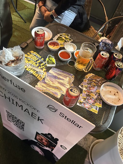A casual gathering with Coca-Cola cans, beer glasses, and various sauces and snacks spread across a metal mesh table, with an 'Interfuse AW 2026 KICKOFF' sign and 'Stellar' and 'SCHIMAEK' logos in the foreground.