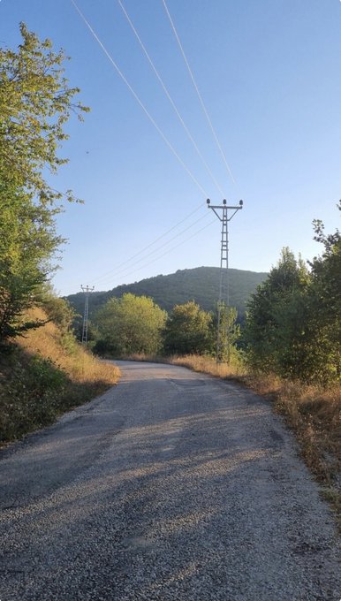 A photograph of a rural asphalt road curving through a hilly, tree-lined landscape under a clear blue sky with visible contrails, featuring an electrical transmission tower on the right side.