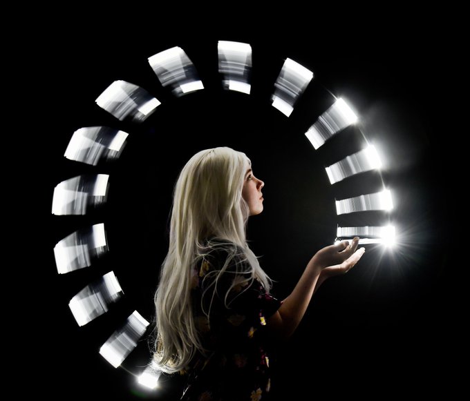 A profile portrait of a long-haired blonde woman holding out her hand toward a circular arrangement of illuminated LED light panels against a black background.