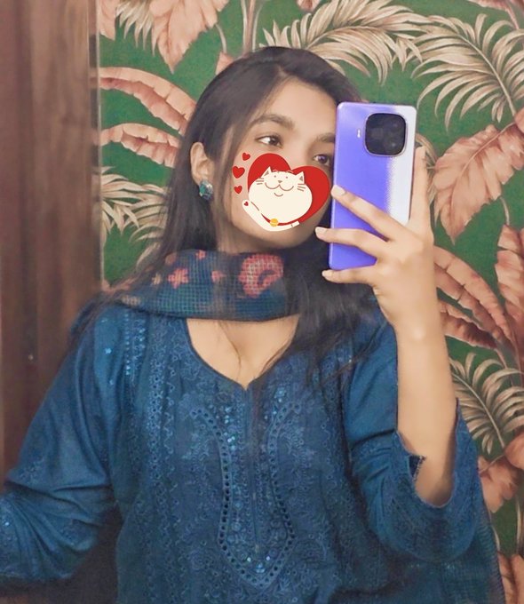 The image features a young woman taking a selfie with her cell phone, holding it up to capture herself in the mirror. She has a cat face sticker on her face, adding a fun and playful element to the photo. The background of the picture is green, which complements the blue dress she's wearing. The caption at the bottom of the image reads "GM Happy Friday & Belated Eid Mubarak Cutiesss Just Wanna say @Polymarket giving me poly trauma :'".
