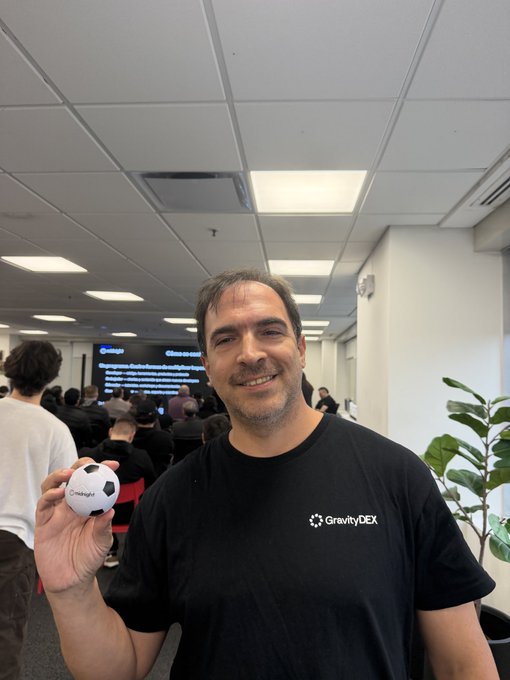 A man in a black 'GravityDEX' t-shirt smiles and holds a soccer ball at what appears to be a tech event or office.
