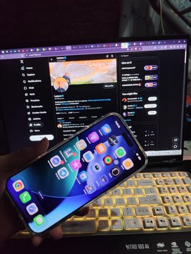 A hand holding an iPhone displaying the home screen over a laptop showing YouTube, with a bottle of "AHA" sparkling water visible.