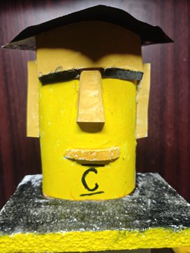 A yellow cylindrical character sculpture with a graduation cap, angry eyebrows, large nose, and mustache, featuring a small black 'C' logo on its chest.