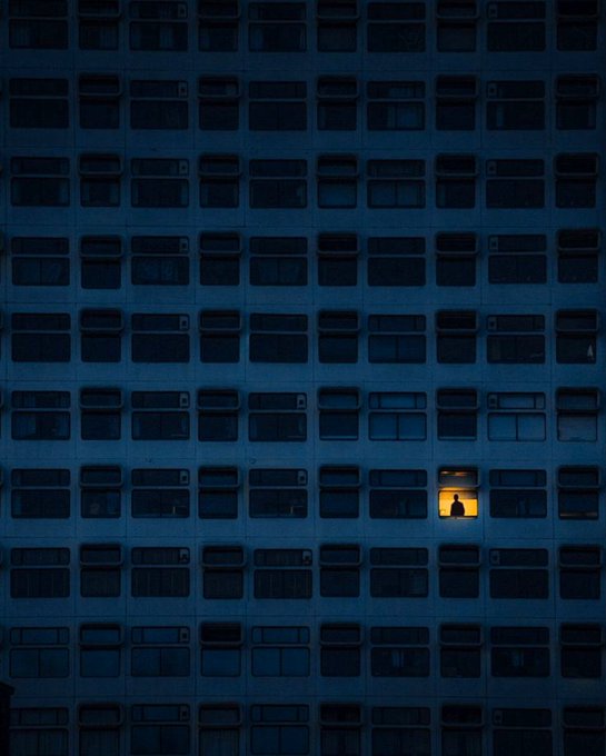 A dark photograph of a large building facade with rows of windows at night, with a single window illuminated in warm yellow light revealing the silhouette of a person standing inside.