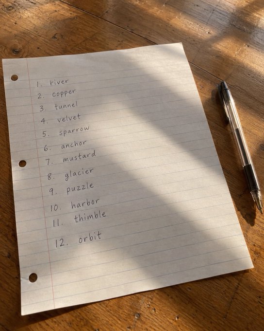 A handwritten numbered list of 12 words (river, copper, funnel, velvet, sparrow, anchor, mustard, glacier, puzzle, harbor, thimble, orbit) on lined paper with a black pen beside it on a wooden surface.