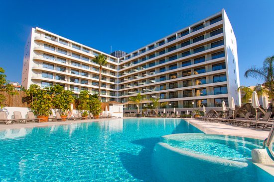 A modern white hotel building with multiple balconies overlooks a bright blue swimming pool surrounded by lounge chairs and palm trees, illustrating the over 2.2 million hotel bookings worldwide now available with XRP through Travala's platform.