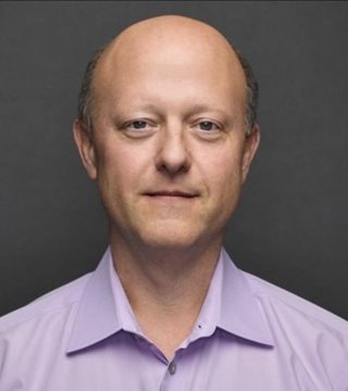 A professional headshot of a bald man wearing a light purple button-down shirt against a gray background.