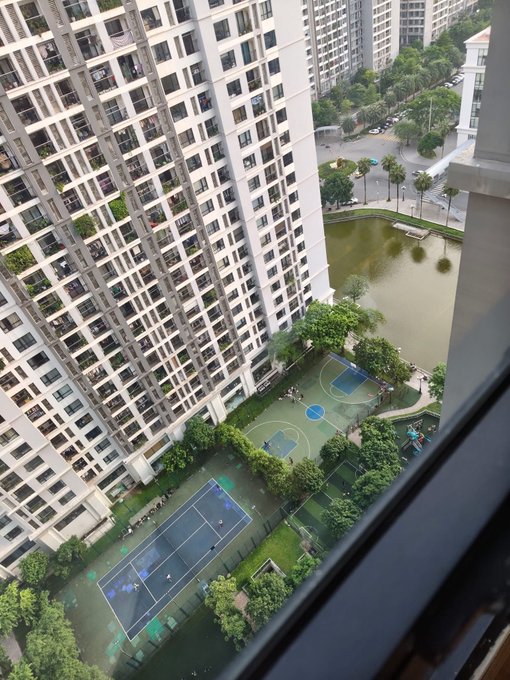 An elevated view from a high-rise apartment window overlooking a residential complex with tennis courts, a basketball court, a pond, and surrounding apartment buildings.