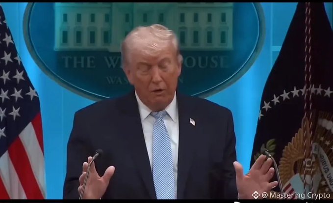The user wants a concise caption for the image.
I need to identify the key elements:
- **Subject:** Donald Trump.
- **Action:** Speaking at a podium, gesturing with hands.
- **Setting:** The White House press briefing room (indicated by the seal behind him).
- **Context from text:** The prompt mentions "TỔNG THỐNG TRUMP" (President Trump) and a quote about Bitcoin and XRP increasing. It also includes a critical comment in Vietnamese about him getting rich from crypto without benefiting others.

**Visual Analysis:**
- The image shows Donald Trump standing at a podium.
- Behind him is the official seal of "THE WHITE HOUSE WASHINGTON".
- To his left is the US flag.
- To his right is the Presidential flag.
- He is wearing a dark suit and a light blue tie.
- His hands are raised in a gesture typical of speaking.

**Synthesizing the Caption:**
The prompt asks