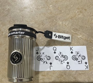 The image features a silver coffee mug with a black lid and handle, as well as a white playing card featuring a black design of a queen of hearts. The mug has the words "Alhamdulillah Just received my merch from @BitgetPak" written on it in black text. The playing card also has the words "Alhamdulillah Just received my merch from @BitgetPak". These items are placed next to each other, suggesting that they may be related or part of a collection.