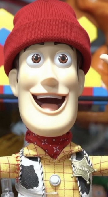 A close-up of a Woody doll from Toy Story wearing his signature red beanie, yellow plaid shirt, cowhide vest, and sheriff badge, with an exaggerated wide-eyed expression.