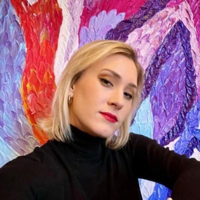 A blonde woman wearing a black turtleneck and red lipstick posing in front of a colorful abstract painting with vibrant red, purple, and blue brushstrokes.