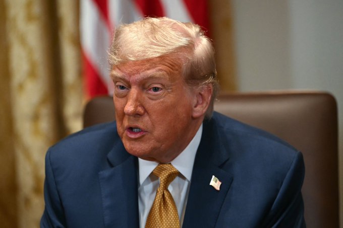 A close-up photo of Donald Trump wearing a dark blue suit and gold tie, with an American flag visible in the background.