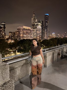 A woman poses on a pedestrian bridge at night with a brightly lit city skyline featuring tall illuminated skyscrapers in the background.