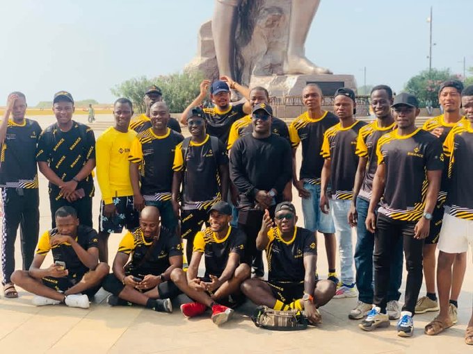 A large group of men wearing matching black and yellow sports jerseys pose for a group photo outdoors in front of a large statue, with some seated in front and others standing behind, in what appears to be a team gathering.