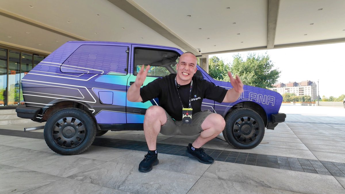 A bald man in a black shirt and shorts squats enthusiastically with arms outstretched in front of a futuristic blue and purple SUV with 'JLRNA' branding.