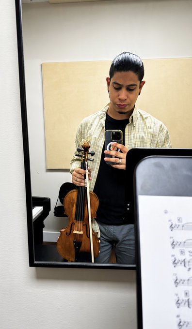 A man takes a mirror selfie while holding a violin and bow, wearing a plaid shirt over a black t-shirt, with a sheet of music visible in the foreground.