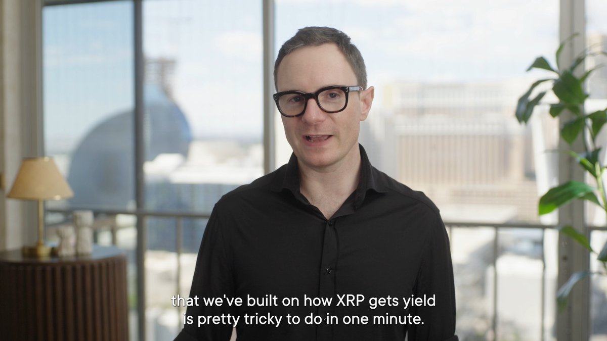 The image shows a man wearing glasses standing in front of a window with a city skyline visible outside. The text above him reads "That we've built on how XRP gets yield is pretty tricky to do in one minute". This suggests that the man might be discussing the intricacies and challenges associated with building and managing an initial coin offering (ICO) like XRP, which can be quite complex due to factors such as token supply, network effects, and market dynamics.