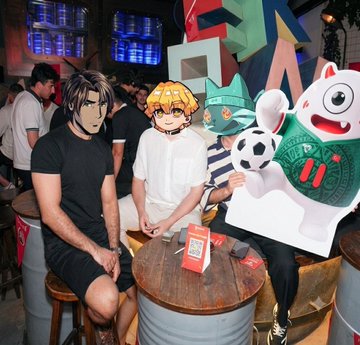 Two people wearing anime character masks (one resembling Uchiha Sasuke, the other a blonde anime character) pose with a soccer-themed mascot at what appears to be a sports bar or fan event, with screens showing above them.