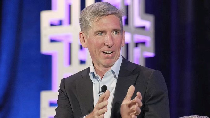 A man in a dark suit and light blue shirt speaks while gesturing with his hands during what appears to be a conference or panel discussion.