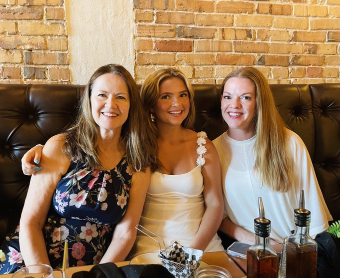 The image shows three women sitting together at a table, smiling for the camera. They are all wearing dresses, with one woman's dress featuring white flowers as an accessory. The setting appears to be indoors, possibly in a restaurant or dining area. On the table, there is a bowl and two bottles of alcohol, suggesting that they might be enjoying drinks while socializing. The post about Bitcoiners reads "3 Generations of Bitcoiners here", which could indicate that these women are part of a family who has been using Bitcoin for an extended period.