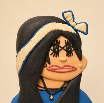 A claymation-style sculpture depicts a female figure with long black hair, large exaggerated lips, thick drawn-on eyelashes, a blue top, and a blue and white striped bow headband.