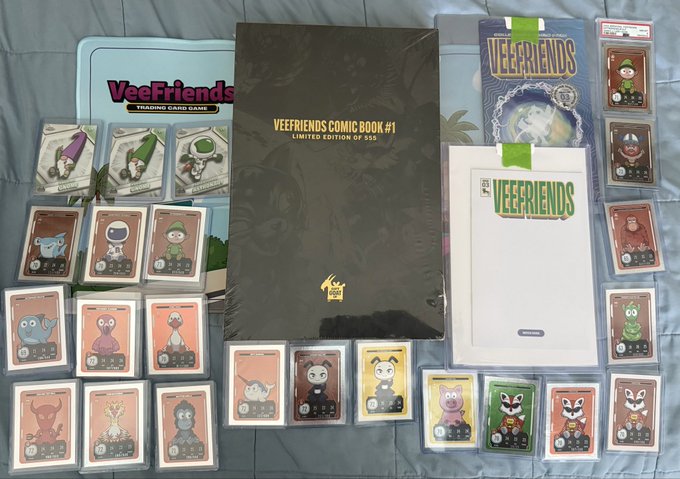 A collection of VeeFriends trading card game merchandise, including a 'VEEFRIENDS COMIC BOOK #1 LIMITED EDITION OF 555' alongside various character trading cards, a trading card game tin, and sealed card packs laid out on a gray surface.