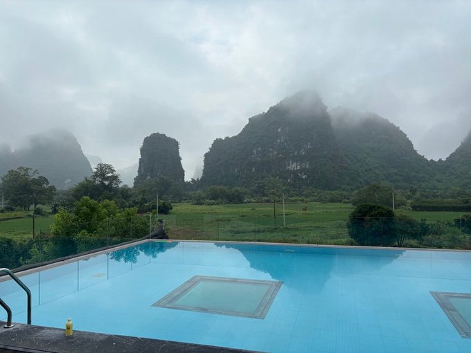 An infinity pool overlooks a misty landscape of lush green karst mountains and rice paddies under a cloudy sky.