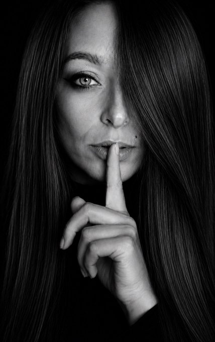 A black-and-white portrait of a woman pressing her finger to her lips in a "shh" gesture, her intense gaze meeting the camera against a dark background — visually echoing the message that silence often speaks louder than words.