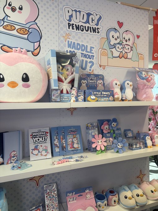 A display shelf features a variety of Puddgy Penguins merchandise, including plush toys, boxed figures, books, and slippers, set against a colorful backdrop reading 'PUDDGY PENGUINS' and 'WADDLE WITHOUT YOU?'