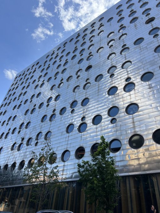 A modern building features a striking silver metallic facade with a distinctive pattern of circular windows of various sizes set against a blue sky with scattered clouds.