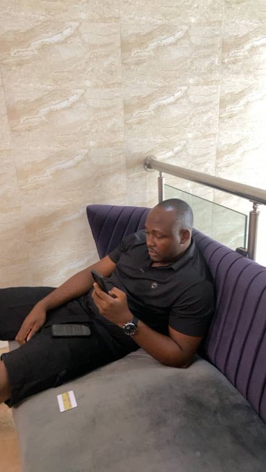A man in a black polo shirt and pants sitting on a gray couch, looking at his phone, with a credit card on the couch beside him.